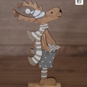 Op de afbeelding: Een houten rendierfiguur met een gestreepte sjaal in wit en bruin, een witte en grijze muts en een grijs en wit gestippeld shirt. Het rendier houdt een kleine zilveren kerstboom vast.