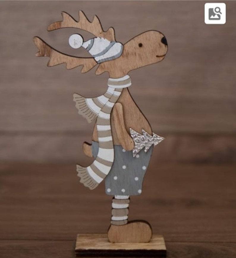 Op de afbeelding: Een houten rendierfiguur met een gestreepte sjaal in wit en bruin, een witte en grijze muts en een grijs en wit gestippeld shirt. Het rendier houdt een kleine zilveren kerstboom vast.