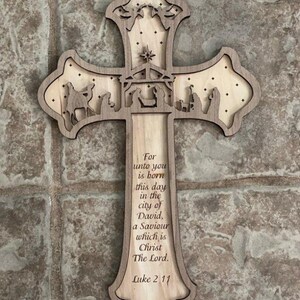 Puede incluir: Cruz de madera con un diseño en capas que presenta una escena de la natividad y el texto "For unto you is born this day in the city of David, a Saviour which is Christ The Lord. Luke 2:11". La cruz es de color marrón claro con vetas de madera natural.