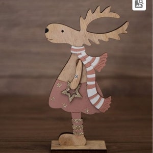 Op de afbeelding: Een houten rendierfiguur met een roze jurk, een wit-roze gestreepte sjaal en een gouden ster. Het rendier staat op een houten voet.