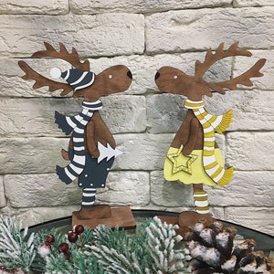 Op de afbeelding: Twee houten rendierfiguren, een met een blauw-wit gestreepte sjaal en een blauwe hoed, de andere met een gele rok en een geel-wit gestreepte sjaal. Beide rendieren staan op houten voetstukken.