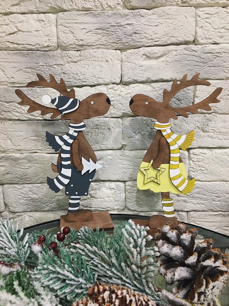 Op de afbeelding: Twee houten rendierfiguren, een met een blauw-wit gestreepte sjaal en een blauwe hoed, de andere met een gele rok en een geel-wit gestreepte sjaal. Beide rendieren staan op houten voetstukken.