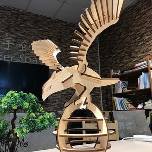 May include: A wooden eagle sculpture with its wings spread, perched on a round, multi-tiered base. The sculpture is made of light brown wood and has a natural finish.