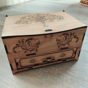 Peut inclure: Une boîte à bijoux en bois avec un tiroir. Le couvercle et le tiroir sont décorés d'un motif de découpe laser d'un arbre aux branches complexes. La boîte est brune avec des accents noirs.