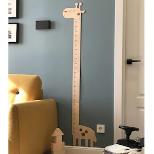 Puede incluir: Un medidor de altura de madera con forma de jirafa montado en una pared azul. El medidor tiene una regla con marcas en centímetros. La jirafa tiene una cara sonriente y manchas. Un sofá amarillo y bloques de madera en el fondo.