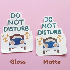 May include: Two stickers with a cartoon design on a pink background. The stickers feature a sleeping blue creature in a bed with the text "DO NOT DISTURB". One sticker has a glossy finish, the other a matte finish.