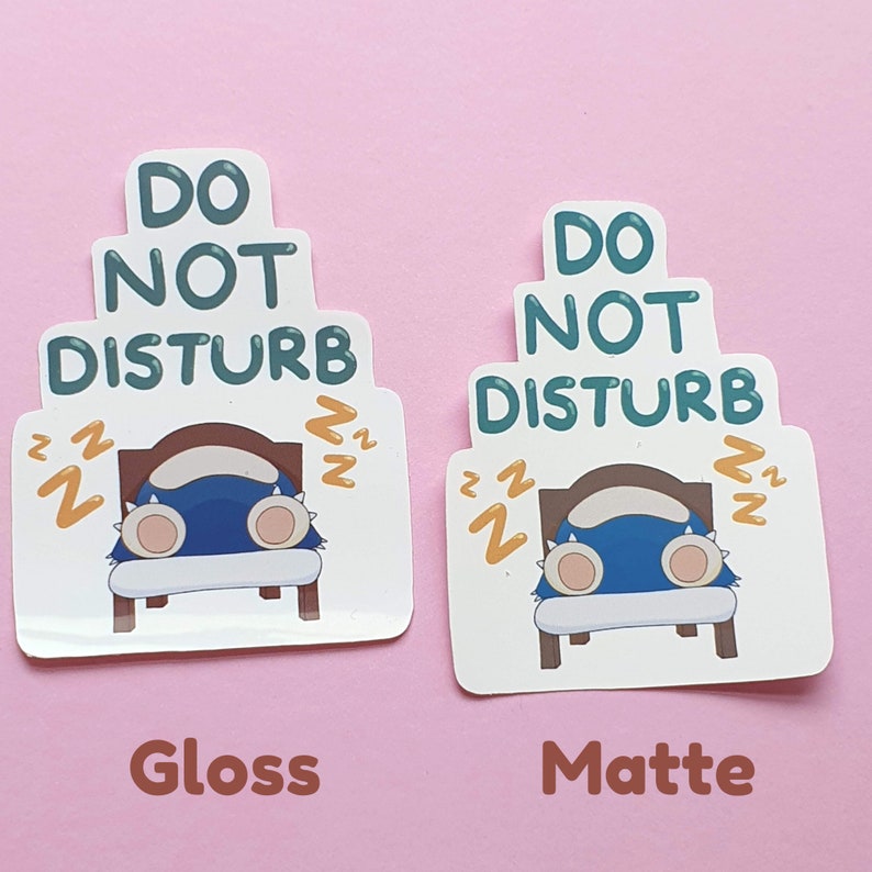 May include: Two stickers with a cartoon design on a pink background. The stickers feature a sleeping blue creature in a bed with the text "DO NOT DISTURB". One sticker has a glossy finish, the other a matte finish.