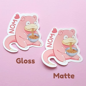May include: Two stickers featuring a pink cartoon character eating noodles from a blue bowl. The character has a long tail and is labeled "NOM" with a heart. One sticker has a glossy finish, the other a matte finish.