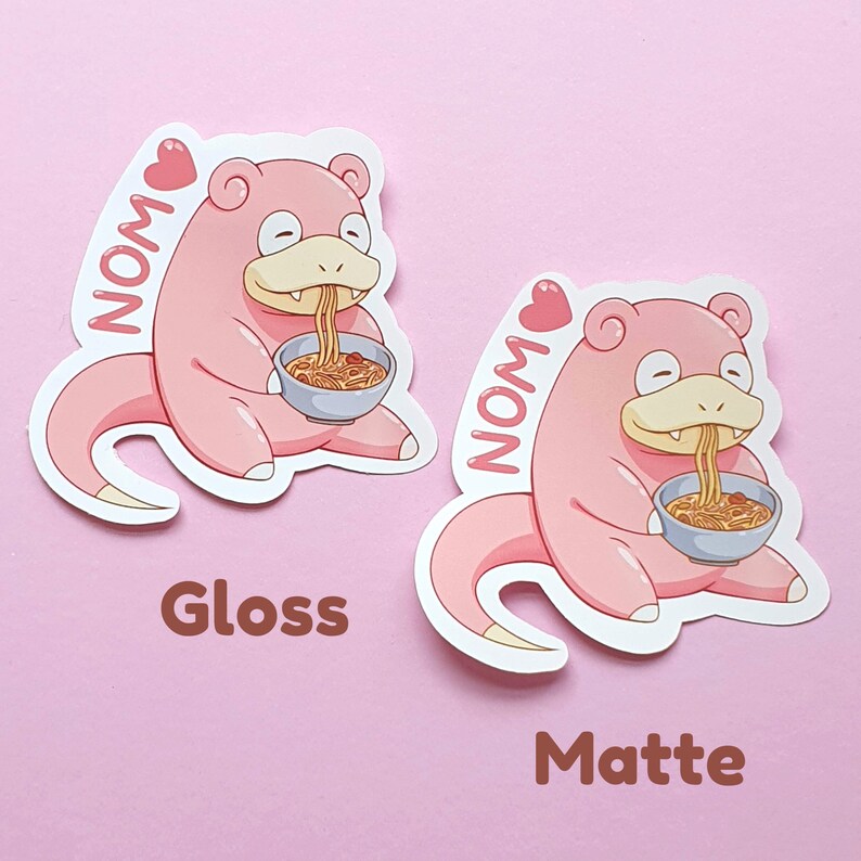 May include: Two stickers featuring a pink cartoon character eating noodles from a blue bowl. The character has a long tail and is labeled "NOM" with a heart. One sticker has a glossy finish, the other a matte finish.