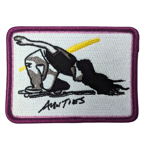 Peut inclure: Écusson brodé avec une bordure violette. Il présente une figure stylisée en position accroupie avec un élément jaune et le texte "AMNTIES" en noir. La figure est représentée en noir, blanc et gris.