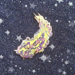 May include: A gold-colored enamel pin with a yellow, pink, and purple design. The pin features a stylized, flowing shape with intricate details. The pin is set against a dark blue fabric with a white star pattern.