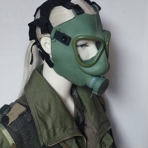 May include: A green gas mask with a black head strap. The mask has a clear visor and a mouthpiece.
