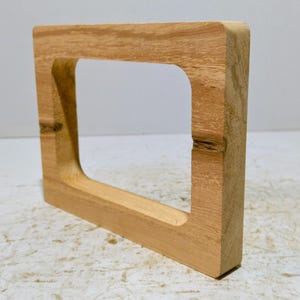 Könnte beinhalten: Ein quadratischer Holzrahmen mit einem rechteckigen Ausschnitt in der Mitte. Der Rahmen besteht aus hellfarbenem Holz mit natürlicher Oberfläche. Die Kanten sind abgerundet, und die Holzmaserung ist sichtbar.