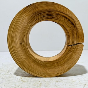 Könnte beinhalten: Rund geformte Holzskulptur mit natürlicher Maserung. Das ringförmige Kunstwerk hat einen Spalt auf einer Seite, wodurch ein offenes Design entsteht. Das Holz hat eine warme, hellbraune Farbe.
