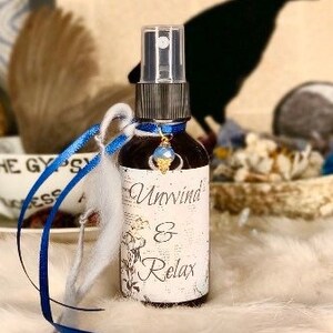 May include: A small glass bottle with a blue and white label that reads "Unwind & Relax". The bottle has a blue and white ribbon tied around it and a blue gemstone charm hanging from the neck of the bottle.