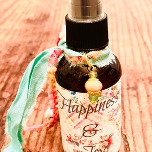 May include: A dark glass spray bottle with a black cap and a label that reads "Happiness & Joy" in script. The bottle is adorned with a light green ribbon, a beaded strand, and a charm. The label features floral designs.