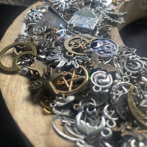 May include: A close-up of a wooden crescent moon-shaped tray filled with a variety of silver and gold charms. The charms are in various shapes and sizes, including stars, moons, and other symbols.