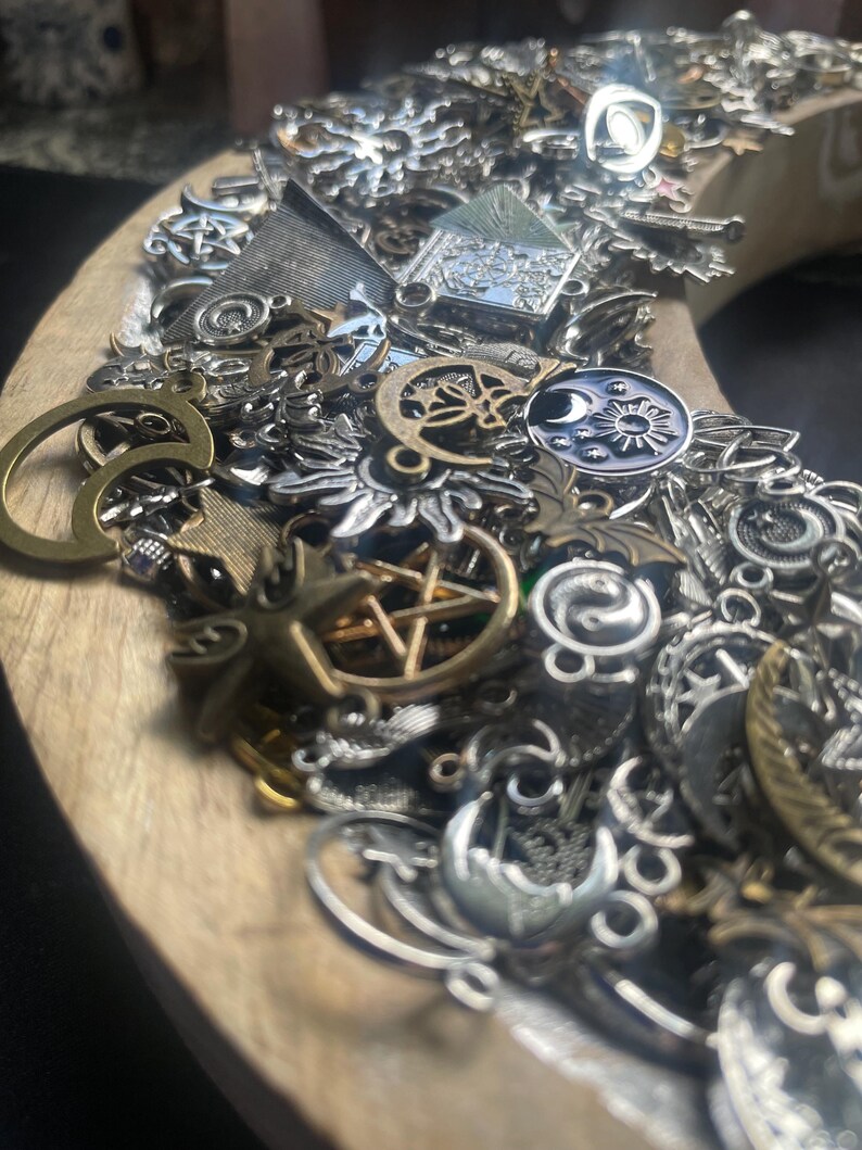 May include: A close-up of a wooden crescent moon-shaped tray filled with a variety of silver and gold charms. The charms are in various shapes and sizes, including stars, moons, and other symbols.