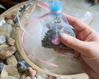 May include: A small, white, sheer bag with a pink ribbon and a silver charm in the shape of two hearts. The bag is filled with dried herbs and flowers. The bag is sitting on a wooden surface with a white background.