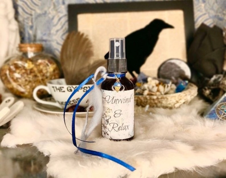 May include: A small glass bottle with a blue and white label that reads "Unwind & Relax". The bottle has a blue and white ribbon tied around it and a blue gemstone charm hanging from the neck of the bottle.