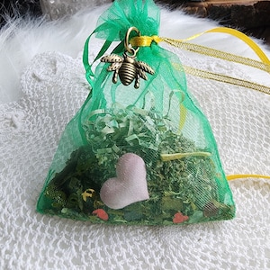 May include: A green mesh bag with a yellow ribbon and a gold bee charm. The bag is filled with dried herbs and a pink heart.