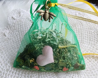 May include: A green mesh bag with a yellow ribbon and a gold bee charm. The bag is filled with dried herbs and a pink heart.