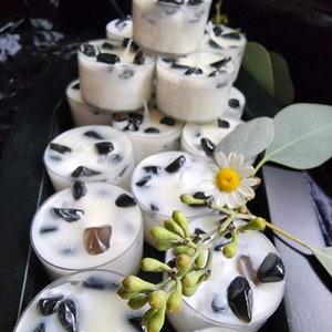 May include: A stack of white tea light candles with black stones embedded in the wax. The candles are arranged in a pyramid shape on a black surface. There are green leaves and a white flower in the background.