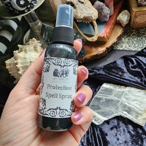 May include: A black glass bottle with a white label that reads "Protection Spell Spray" and "Life Spirits" in a decorative font. The bottle is in a hand with purple nail polish.