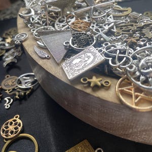 May include: A wooden bowl filled with a variety of silver and gold charms, including stars, crosses, moons, and other symbols. The charms are arranged in a random pattern and some are overlapping.