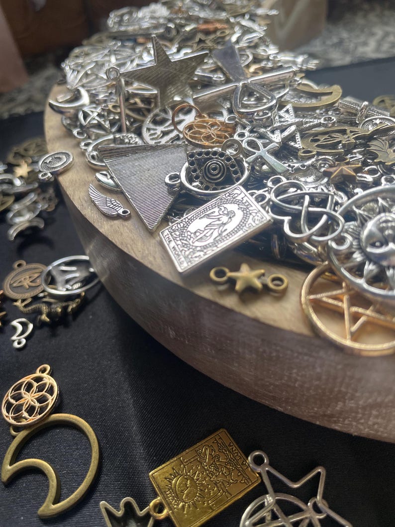 May include: A wooden bowl filled with a variety of silver and gold charms, including stars, crosses, moons, and other symbols. The charms are arranged in a random pattern and some are overlapping.