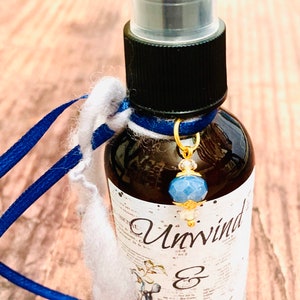 May include: A small brown glass bottle with a black spray top and a blue and gold charm. The bottle is labelled "Unwind & Relax".