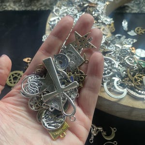 May include: A hand holding a variety of silver charms, including a star, a cross, a lotus flower, and a crescent moon. The charms are all attached to a chain and are likely for jewelry making.