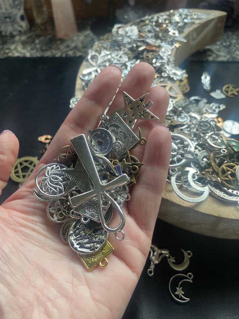 May include: A hand holding a variety of silver charms, including a star, a cross, a lotus flower, and a crescent moon. The charms are all attached to a chain and are likely for jewelry making.