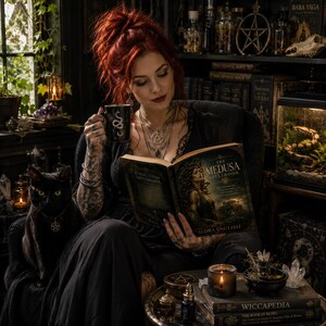 May include: A person with red hair sits in a chair holding a black mug with a white snake graphic and reading a book titled The Medusa Spellbook by Audra English. A black cat with a pentagram collar sits beside them in a room filled with books.