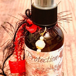 May include: A brown glass bottle with a black spray top and a label that says "Protection & Evil Eye". The bottle is decorated with red and black feathers and a gold charm.