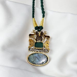 May include: A gold-toned pendant necklace with a green cord. The pendant features a rectangular green stone, an oval blue stone, and decorative elements including deer and faces. The necklace is displayed on a white background.