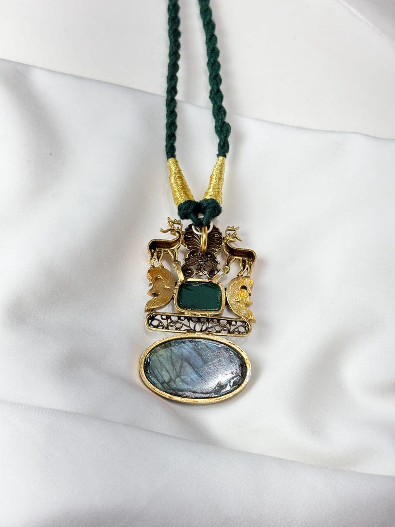 May include: A gold-toned pendant necklace with a green cord. The pendant features a rectangular green stone, an oval blue stone, and decorative elements including deer and faces. The necklace is displayed on a white background.