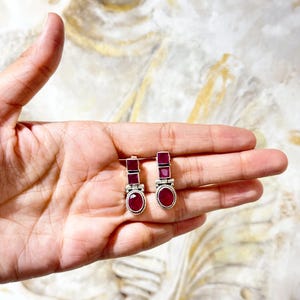 May include: A pair of silver-toned earrings with rectangular and oval ruby-colored gemstones. The earrings are held in a hand, showcasing the contrast between the silver metal and the deep red of the stones. The earrings have a classic, elegant design.