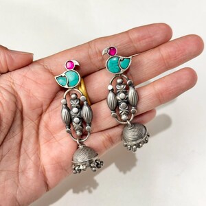May include: A pair of silver-toned earrings with a peacock design. Each earring features a turquoise body, a pink head, and a dangling bell-shaped bottom. The earrings are displayed on a light background.