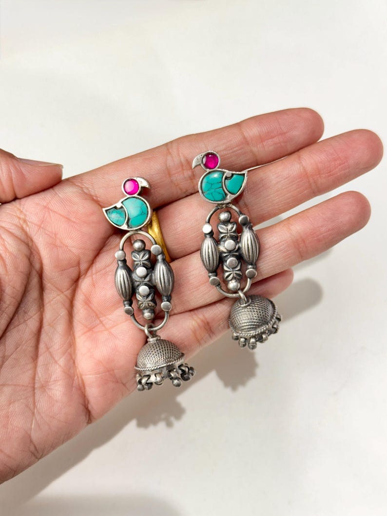 May include: A pair of silver-toned earrings with a peacock design. Each earring features a turquoise body, a pink head, and a dangling bell-shaped bottom. The earrings are displayed on a light background.