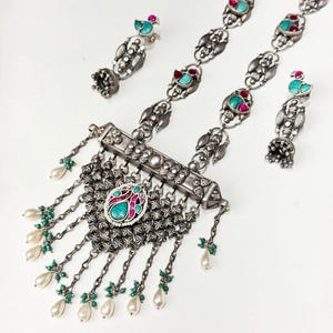 May include: A silver-toned jewelry set featuring a necklace and matching earrings. The necklace has a central pendant with turquoise and pink accents, dangling pearl and green bead strands. The earrings have a bird design with turquoise and red accents.
