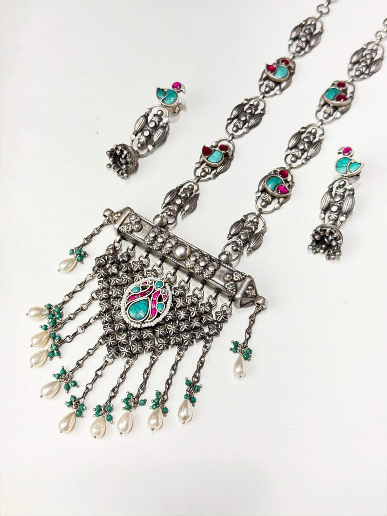 May include: A silver-toned jewelry set featuring a necklace and matching earrings. The necklace has a central pendant with turquoise and pink accents, dangling pearl and green bead strands. The earrings have a bird design with turquoise and red accents.