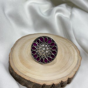 May include: A silver ring with a round design featuring a central flower motif surrounded by purple enamel petals.