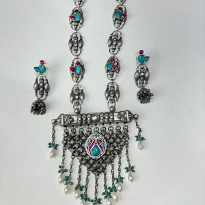 May include: A silver-toned necklace and earring set. The necklace features a central pendant with turquoise and pink accents, and dangling pearl-like beads. The earrings have matching turquoise and pink details. The set is ornate and detailed.
