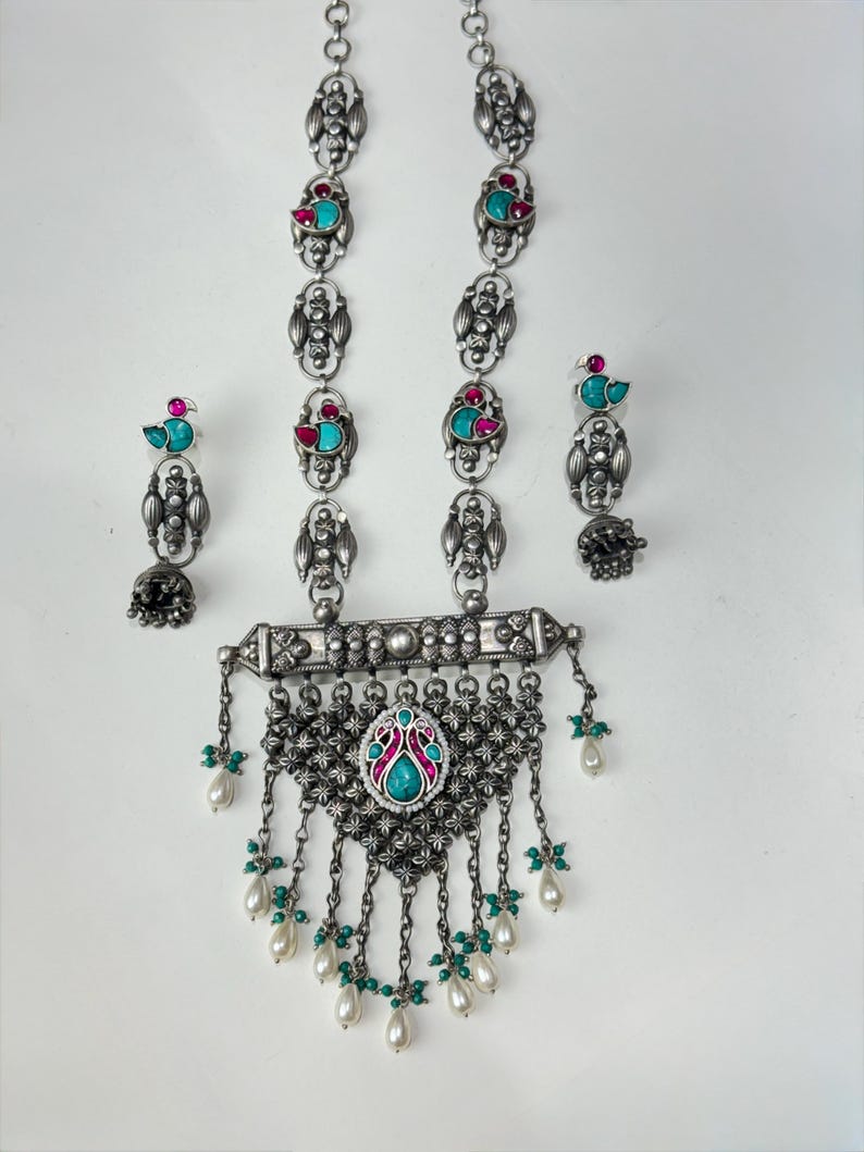 May include: A silver-toned necklace and earring set. The necklace features a central pendant with turquoise and pink accents, and dangling pearl-like beads. The earrings have matching turquoise and pink details. The set is ornate and detailed.