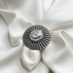 May include: A silver ring featuring an intricate design of a face in the centre. The ring is encircled by a textured, circular band.