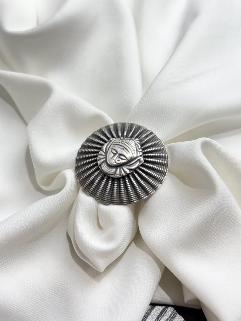 May include: A silver ring featuring an intricate design of a face in the centre. The ring is encircled by a textured, circular band.