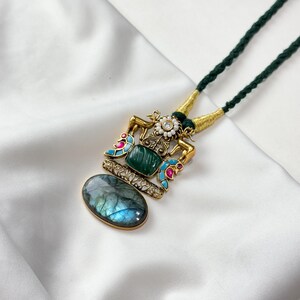 May include: A gold-toned pendant necklace with a green cord. The pendant features a central green stone, a blue-gray oval stone, and colorful enamel accents. The necklace is displayed on a white, textured surface.