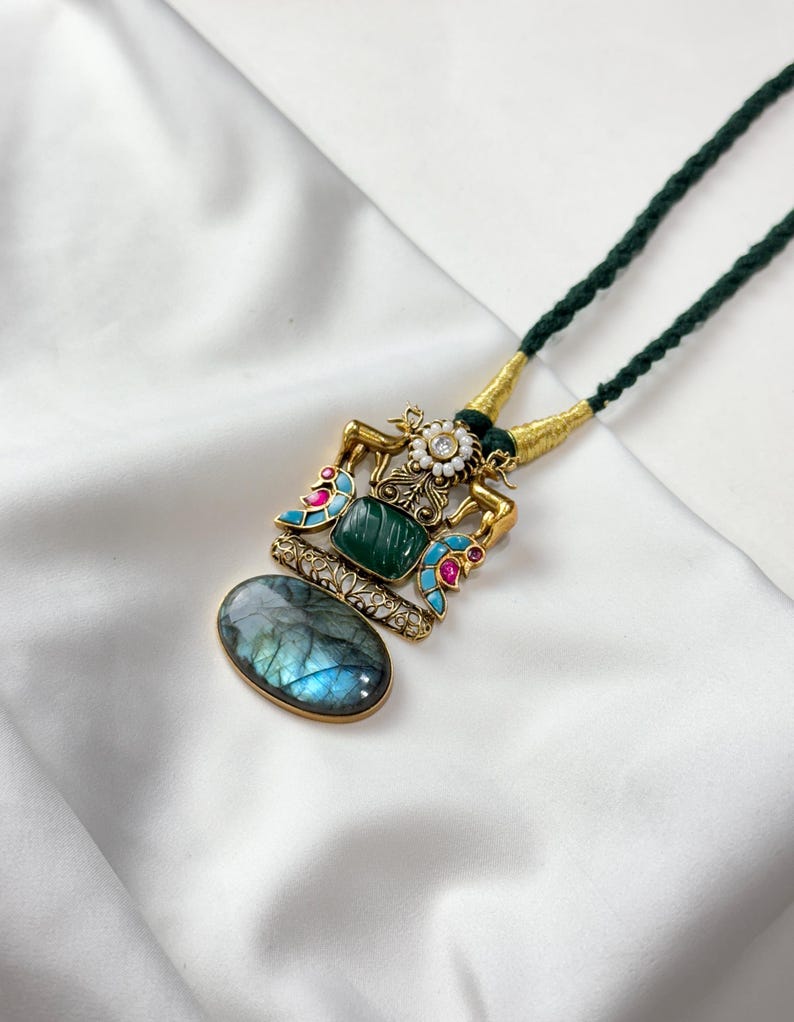 May include: A gold-toned pendant necklace with a green cord. The pendant features a central green stone, a blue-gray oval stone, and colorful enamel accents. The necklace is displayed on a white, textured surface.