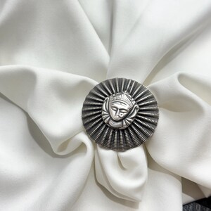 May include: A silver ring with a circular design featuring a stylised face in the centre. The ring is on a white fabric background.